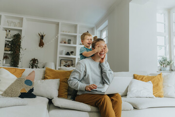 Smiling boy covering mother eyes while standing on sofa at home