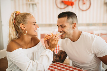 Cheerful young woman feeding pizza to boyfriend while sitting in restaurant