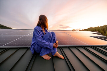 Rooftop with solar panels on house in mountains, woman sitting alone raising hands and enjoying sunset. Energy independence, sustainability, self sufficient, and escapism to nature concept