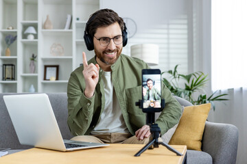 Man wearing headphones and using his smartphone and laptop for recording a video at home. He is smiling and pointing upwards.