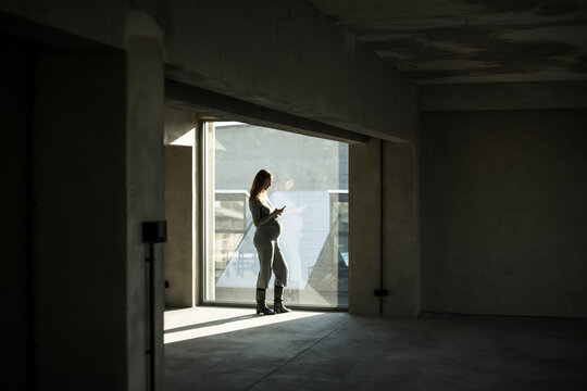 Pregnant businesswoman standing at office window, using smartphone - Powered by Adobe
