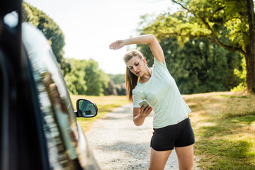 Sportive young woman stretching and using cell phone at a car in a park