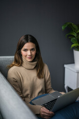 Portrait of content woman wearing light brown turtleneck pullover sitting on couch with laptop