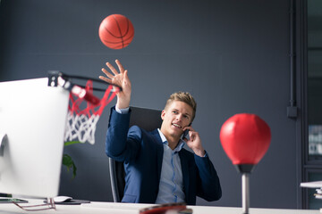 Young businessman sitting at desk in office playing basketball and talking on cell phone