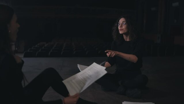 A confident and interested brunette girl with curly hair in round glasses and a black theatrical costume communicates with her colleague and they play roles preparing to study for a performance on