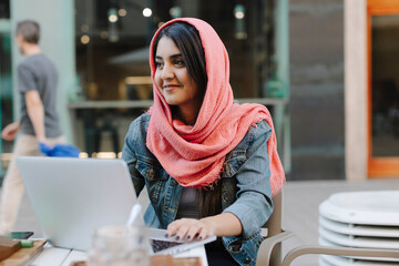 Smiling young woman with laptop wearing headscarf at a pavement cafe - Powered by Adobe