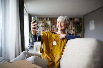 Smiling senior woman taking selfie with new blouse on smart phone while sitting at home