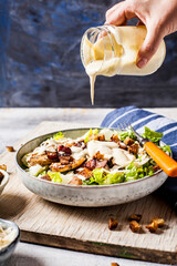 Hand of person pouring dressing over bowl of Caesar salad with romaine lettuce, Parmesancheese, bacon, chicken breast and croutons