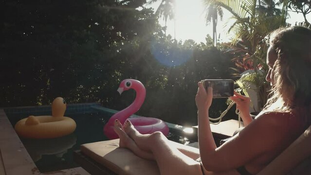 Woman in bikini relaxes near swimming pool in Thailand resort, make photo on phone, flamingo and duck inflatable rings float nearby. Girl relax outdoor travel summer holiday vacation in private villa