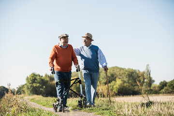 Old friends taking a stroll in the fields with walking stick and wheeled walker, talking about old...
