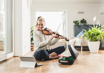 Woman with tablet sitting on the floor at home playing violin