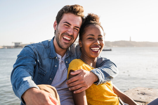 Portrait of happy young couple sitting at the waterfront, Lisbon, Portugal