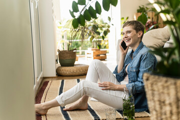 Cheerful woman talking over smart phone while sitting on carpet at home
