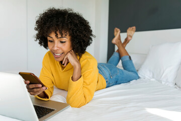 Smiling woman lying on bed using cell phone and laptop