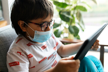 Boy with surgical mask sitting on armchair using digital tablet