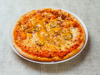 A pizza with cheese and pickles on top. Italian dish with dough, sauce and cheese. Simple white table cloth. Popular food.