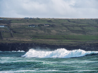 A rocky shoreline with a large wave crashing against the rocks. The sky is cloudy and the water is choppy. Cliff in the background. Doolin area, Ireland. Popular tourist area.