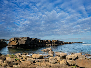 A rocky shoreline with a cloudy sky in the background. The rocks are scattered along the shoreline, and the water is calm. Scene is peaceful and serene, with the natural beauty of the rocky shoreline