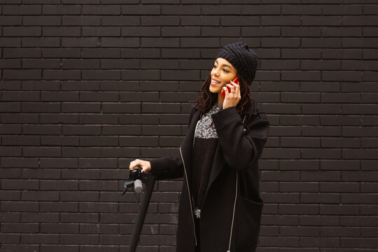 Young woman with dreadlocks smiling while talking on a red smartphone on an electric scooter, standing against a black brick wall.