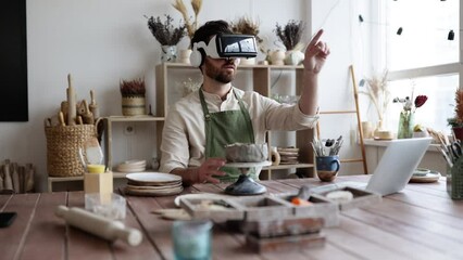 Sculptor in pottery studio uses virtual reality glasses to sculpt vase from clay. Workspace filled with pottery tools and materials. Blend of traditional craft with technology show innovation in art.