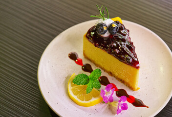 Close up a blueberry cheesecake with cute decoration in a white plate with a sliced of lemon. Small thin gold sheets on the blueberries. View on above, black table with texture surface.