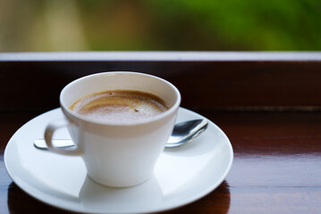 Close up a white cup of hot coffee on wooden table with blurred background of nature, space for copy, natural morning light.