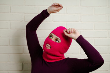 A woman wearing a bright pink balaclava dances energetically, striking unique poses with her hands...