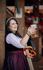 Young, beautiful Bavarian woman toasting with beer in crowd and holding a big pretzel