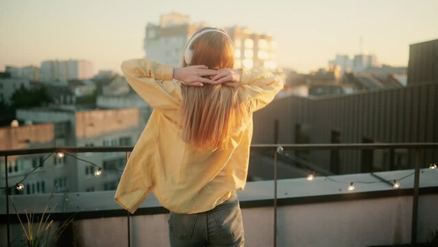 Young excited blonde woman in earphones dancing outside on rooftop. Slow motion of happy girl in wireless headphones dancing singing on a Roof at Sunset having fun alone