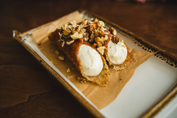 Two cannoli on a plate with. The cannoli are filled with cream and topped with nuts. The plate is a simple white ceramic plate with a brown border.