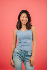Vertical. Teenage asian female student looking at camera and standing at red studio background. Young adult chinese woman staring front at pink colour backdrop. Asiatic girl with friendly expression