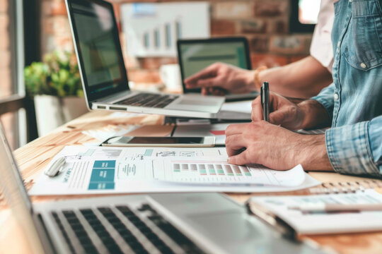 Professional designers collaborating over graphic designs on laptops in a vibrant, sunlit workspace filled with creative tools.