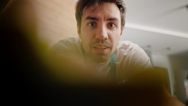 Confident brunette guy with stubble in a white T-shirt and blue apron wipes the camera with a yellow rag and poses with it while cleaning the kitchen in a modern apartment