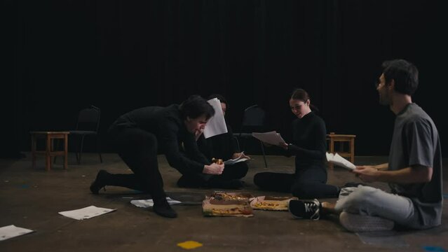 A trio of confident young actors in black suits chat about the script and eat pizza during a rehearsal for a new play in a theater on stage. Happy actors eat pizza during a break during a play