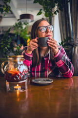 A woman sits at a cafe table and enjoys a cup of tea. The cafe is bright and airy, with windows letting in natural light. The woman wears glasses and a pink and black plaid shirt.