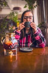 A woman sits at a cafe table and enjoys a cup of tea. The cafe is bright and airy, with windows letting in natural light. The woman wears glasses and a pink and black plaid shirt.