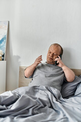 A man with inclusivity lies in bed, covered by a blanket, talking on a cell phone.