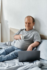 A man with inclusivity sits in bed, enjoying a cup of coffee while working on his laptop.