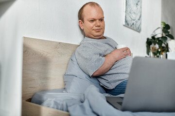 A man with inclusivity sits in bed, enjoying a cup of coffee and working on his laptop.