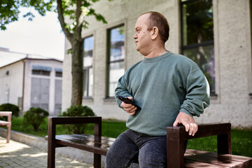 Fototapeta premium A man with inclusivity sits on a bench, holding a phone, in front of a building.