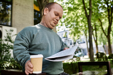 Fototapeta premium A man with inclusivity sits outdoors on a bench, reading a newspaper and enjoying a cup of coffee.