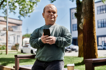 Fototapeta premium A man with inclusivity sits on a bench in a city park, using his smartphone while enjoying a cup of coffee.