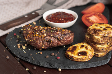 Roasted pork steak on black stone board over dark background. With tomato, sauce and fried potato