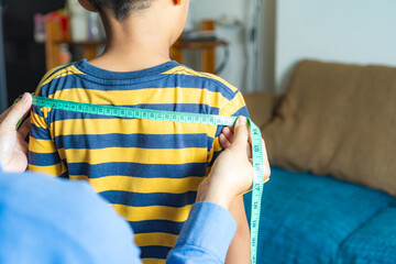 A mother measures her child's shoulder width using a measuring tape at home, concept of shopping for children's clothes online