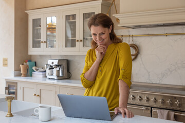 Happy senior woman at home in her kitchen using a laptop to look at his pension fund