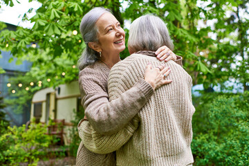 Two women, wearing cardigans, hug each other in a lush green setting. They appear to be a couple, enjoying a moment of affection outdoors.