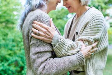 A middle-aged lesbian couple embraces each other warmly while wearing cardigans, surrounded by lush...