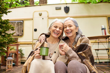 Two middle-aged women, a lesbian couple, laugh together while sitting in front of their camper van in a lush forest setting.
