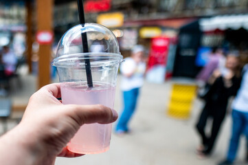 A hand holds a clear plastic cup with a pink beverage and a dome lid, featuring a straw, against a blurred street background