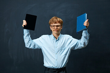 Young man with red hair and glasses stands holding tablet in his right hand and blue book in his...